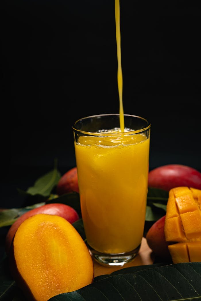 Vibrant fresh mango juice pouring into glass surrounded by ripe mangoes and leaves.